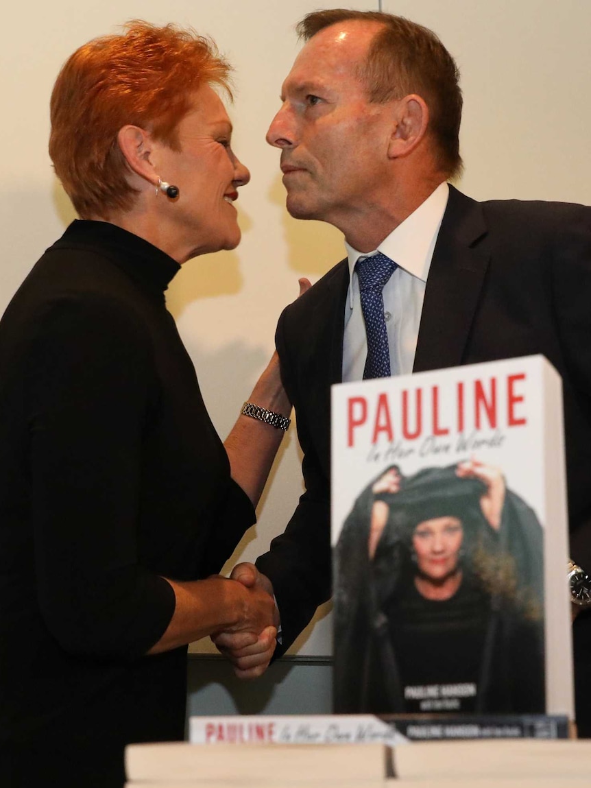 Tony Abbott and Pauline Hanson shake hands behind a copy of a the One Nation leader's book