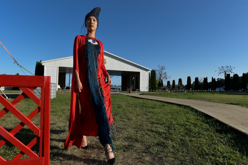 A model wearing designer clothes stands in a field with a shelter behind her. 