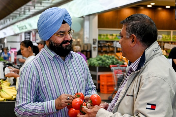 A man wearing a turban and holding some tomatoes talks to another man in a fresh food market.