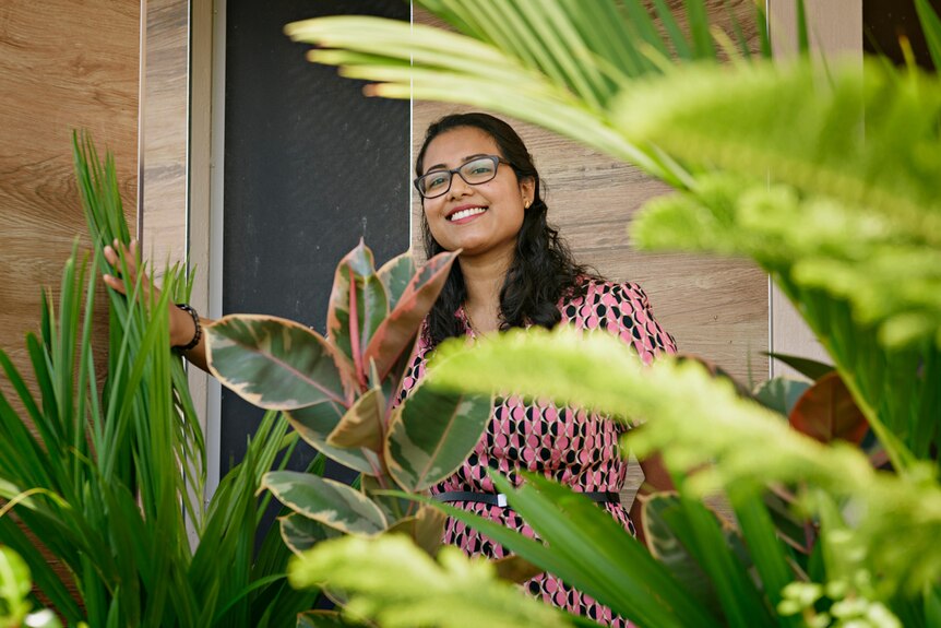 A woman with black hair and brown skin smiles among green shrubbery