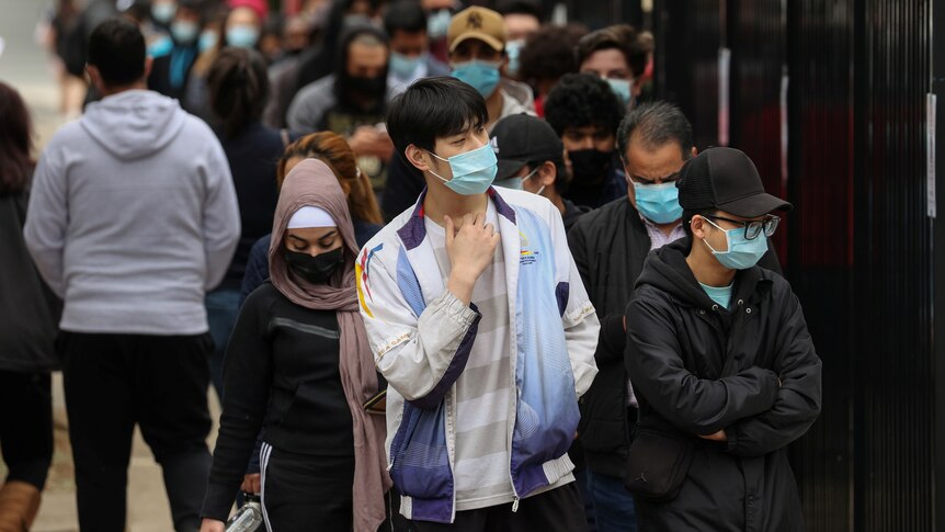 people wearing masks stand in a queue along a fenceline  