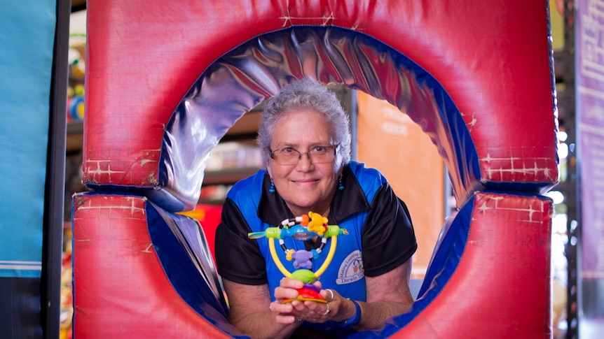 A woman lies in a rubberised toy tunnel holding a plastic baby's toy.