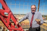 A man in a grey shirt and red tie stands on a crane holding a gold bar over the city skyline.