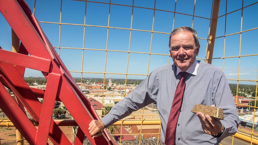 A man in a grey shirt and red tie stands on a crane holding a gold bar over the city skyline.