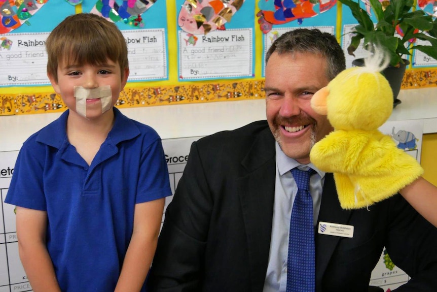 A student wearing a blue shirt and tape as a moustache sits next to school principal Anthony Middleton and a duck hand puppet.