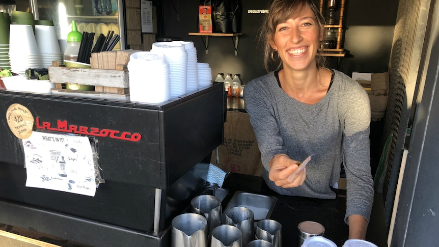 Woman standing behind coffee machine with takeaway coffees smiling, holding change.
