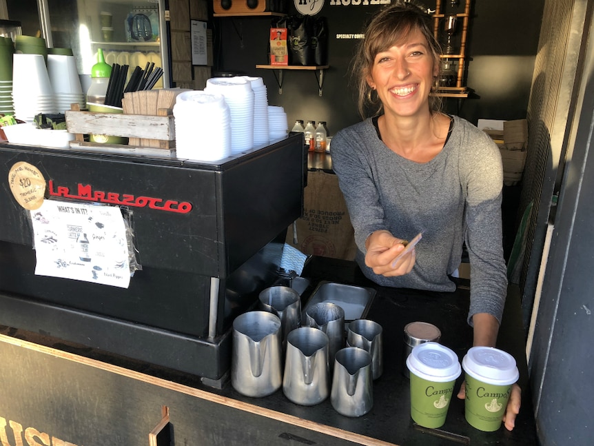 Woman standing behind coffee machine with takeaway coffees smiling, holding change.