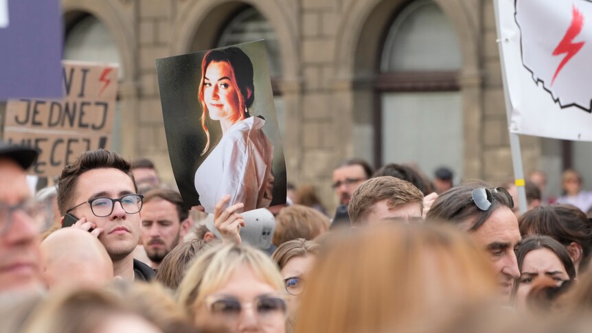 A large crowd a gathers for a protest in daylight, with someone holding aloft a photo of smiling young woman.