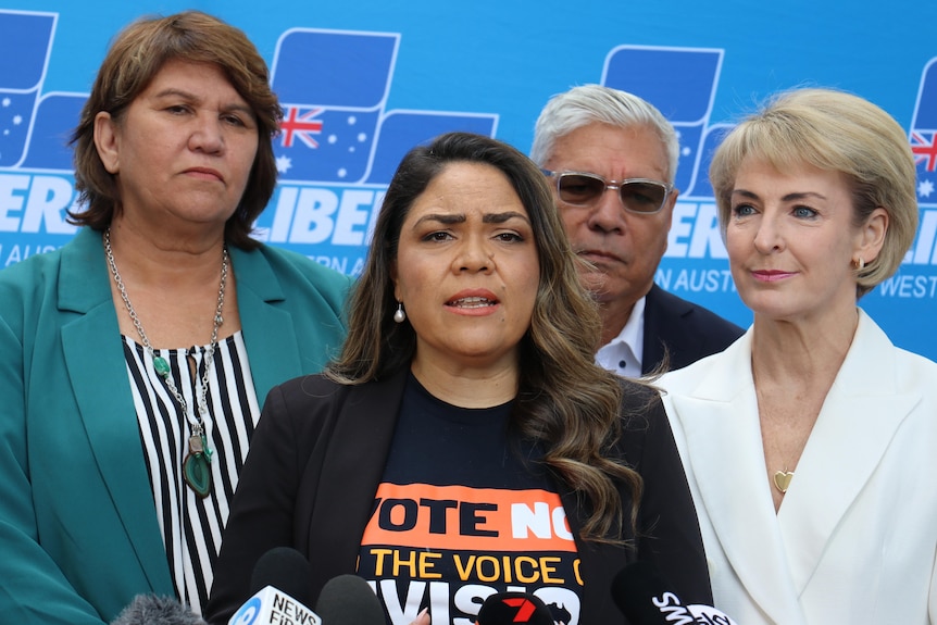 An Indigenous woman a suit speaks to journalists, while three women stand behind him. There's a Liberal WA backdrop.