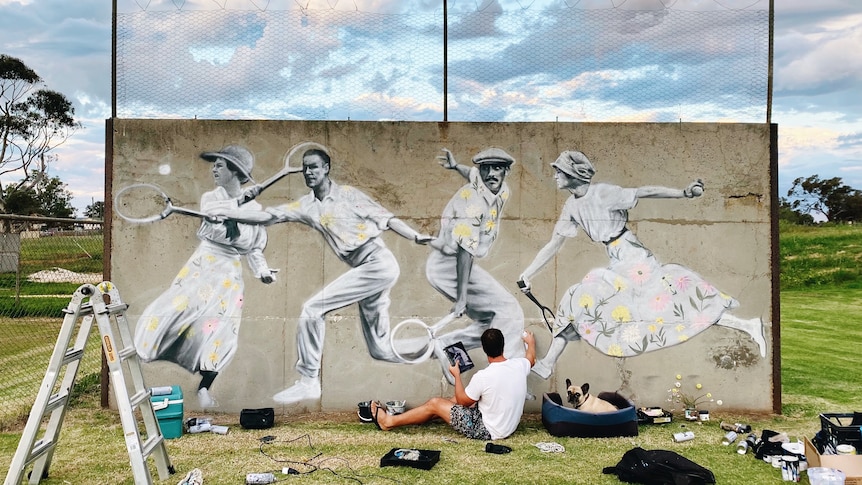 A man in shorts painting a mural of old fashioned tennis players on a faded concrete wall. His dog sits beside him on a cushion.