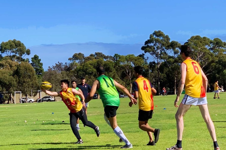 A young and small Asian man stretches for an Aussie Rules ball surrounded by taller teammates in matching yellow shirts.