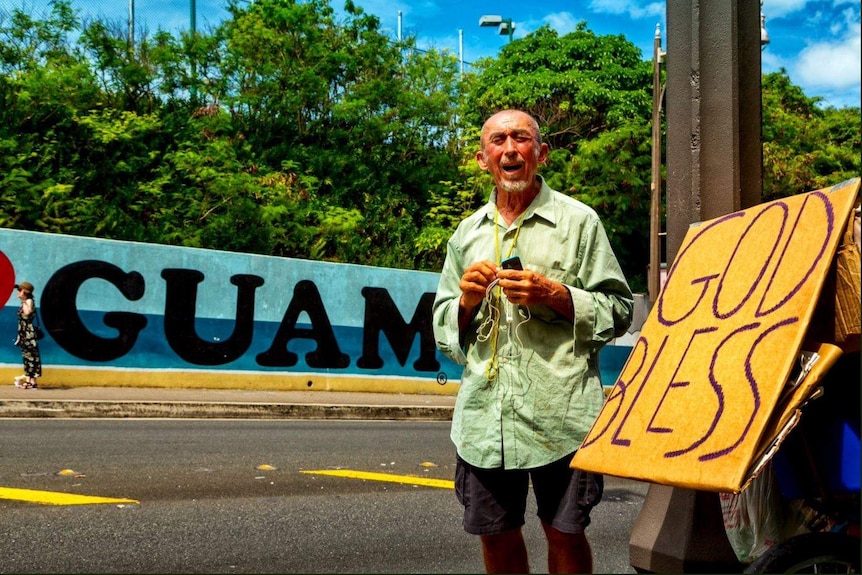 A man looks at the camera with a sign in the foreground saying "God bless" and one in the background saying "Guam".