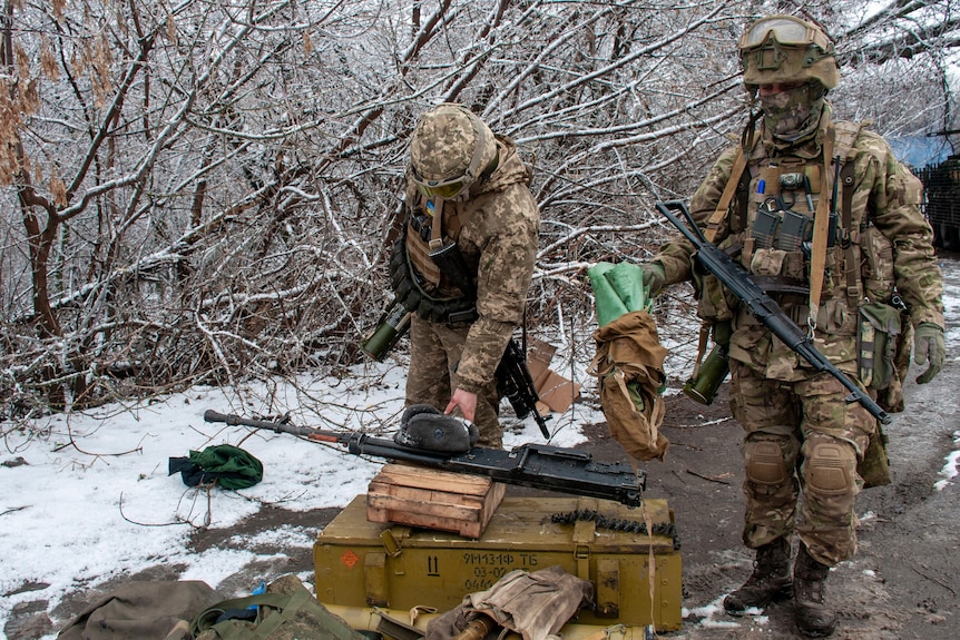 Soldiers in battle gear ready their rifles at a snowy defensive position behind leafless trees.