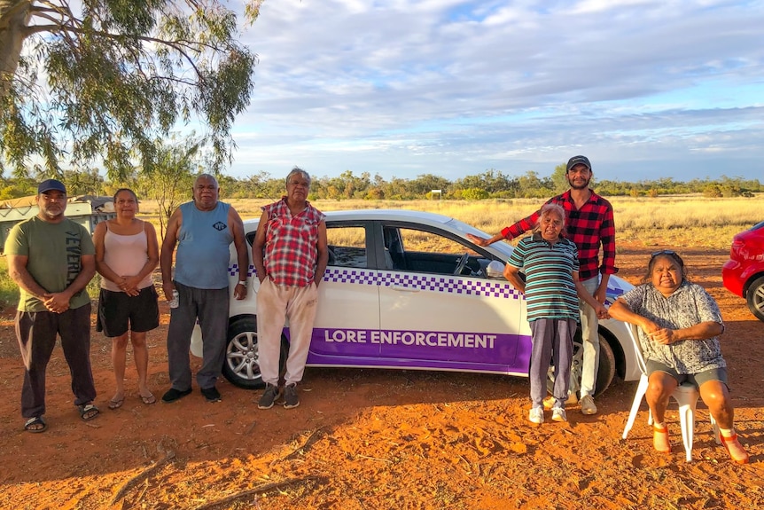 Seven people including some Indigenous elders standing or sitting on red dirt next to a car labelled 'Lore Enforcement'