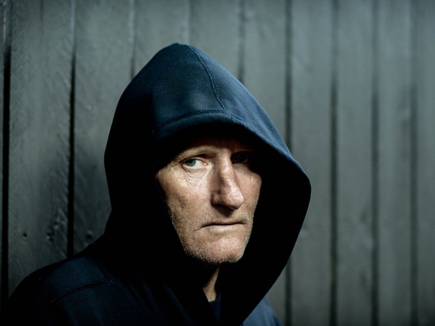 A middle-aged man in a dark hoodie looks into camera with a serious expression.