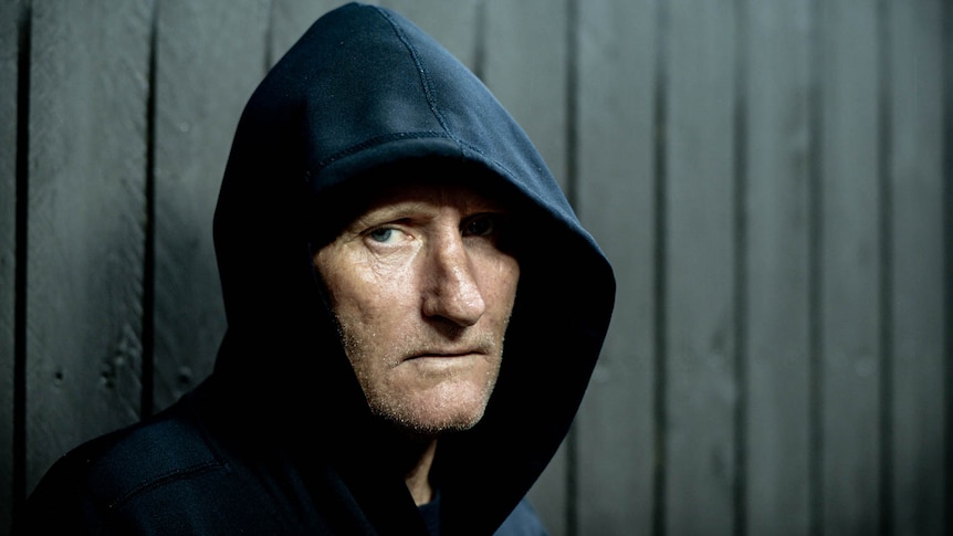 A middle-aged man in a dark hoodie looks into camera with a serious expression.
