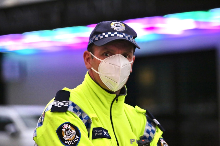 A police officer wearing a fluoro yellow jacket and white face mask stares into the camera intently.