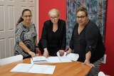 Three professional women stand around a table looking at documents