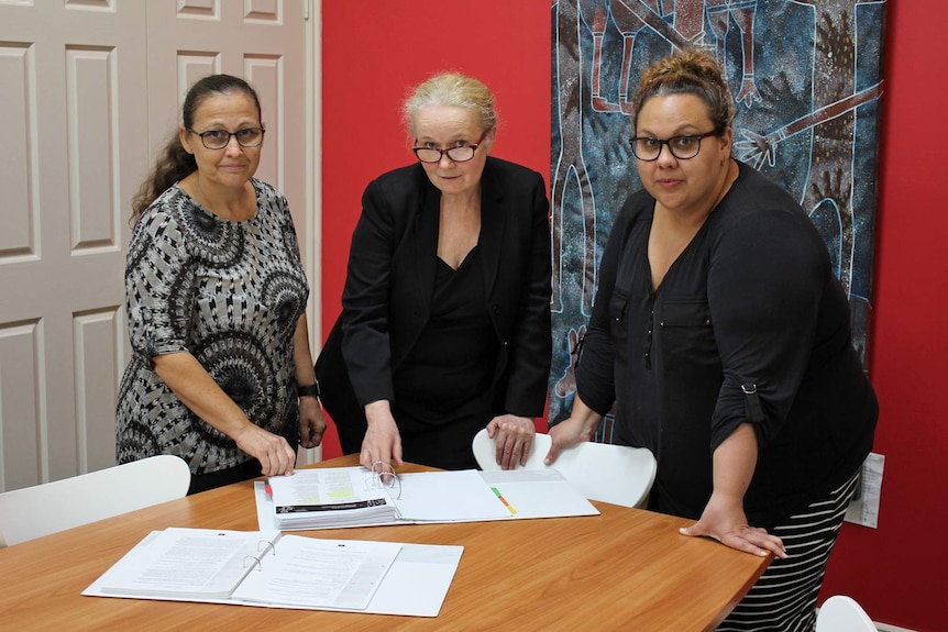 Three professional women stand around a table looking at documents