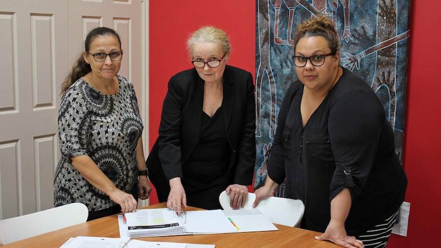 Three professional women stand around a table looking at documents
