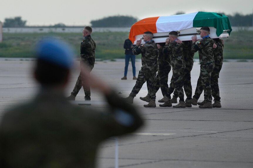 A small group of soldiers carry a white coffin draped with an Irish flag as another soldier salutes.