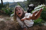 White woman with brown hair screams and reaches for help on a forest floor while a large bear approaches from behind her.