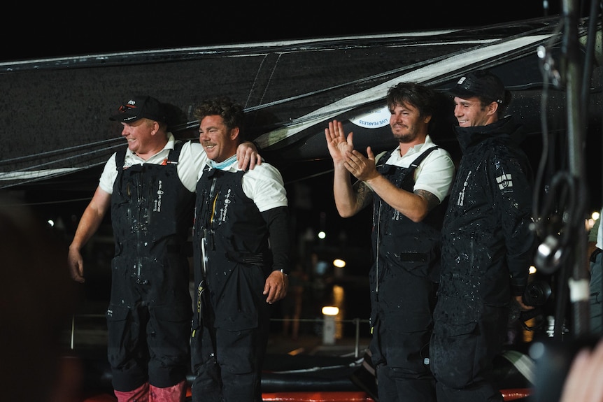 Four men in black overalls stand on a yacht with arms around each other or clapping.