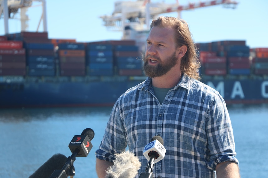 David Knoff wearing a blue checked long sleeve shirt, standing in front of microphones with ships in the background.