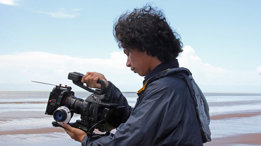 Sydney-based filmmaker Nathaniel Kelly stands with a camera pointing out towards Casuarina beach in Darwin.