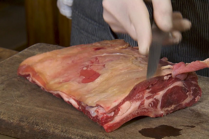 A slab of goat meat on a chopping board with a knife slicing through the meat.