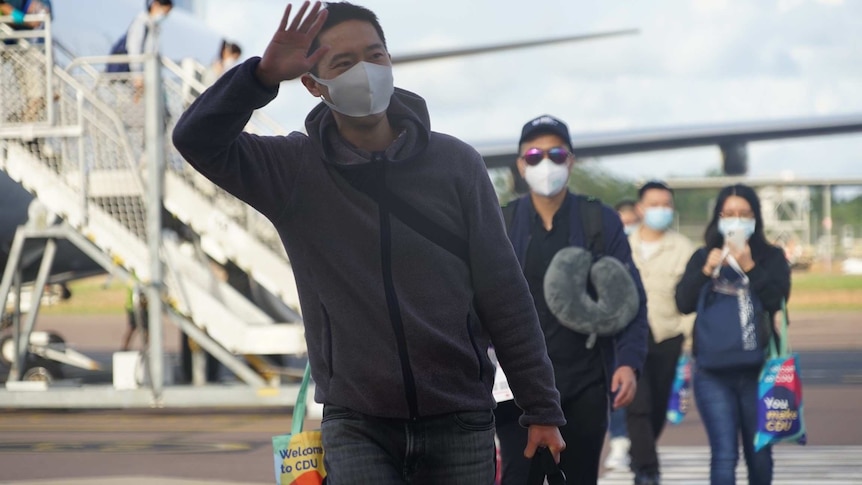 A student wearing a face mask waves on the Darwin Airport tarmac with other students walking behind him.