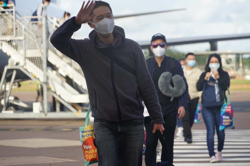 A student wearing a face mask waves on the Darwin Airport tarmac with other students walking behind him.