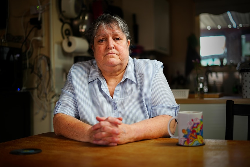 A woman with short grey hair sitting at her dining table with a mug. 