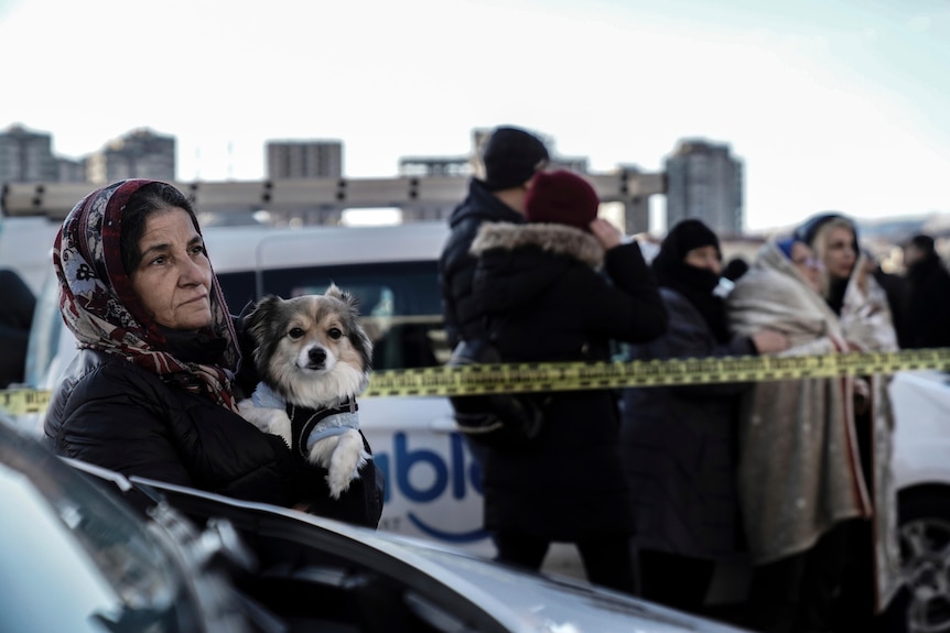 A woman holding a dog watches, the emergency teams as they search for survivors in the rubble of a destroyed building.