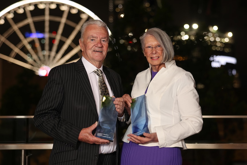 A man and woman with grey hair each hold a glass award, with a lit up ferris wheel in the background.