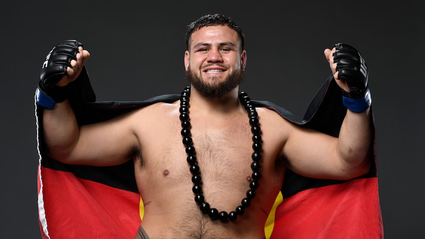 Tai Tuivasa clenches his fists, smiles, and holds up an Aboriginal flag wearing a Samoan necklace