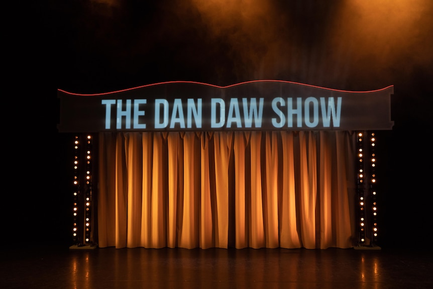 An empty stage with closed curtains bathed in orange light, with a sign reading The Dan Daw Show.