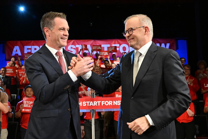 a man shaking and holding the hand of another man who is wearing glasses as they both stand in front of a campaign meeting
