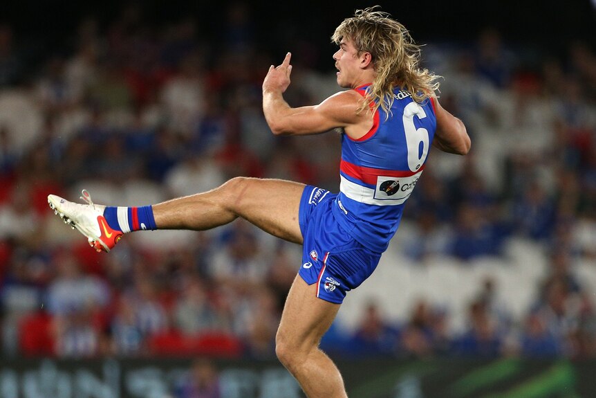 An AFL player with a mullet kicks into the air.