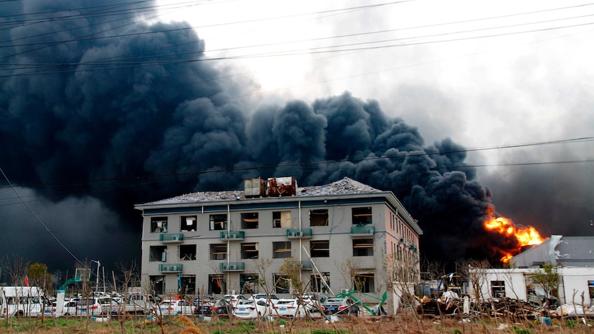 Dark black smoke billows from a burning fire near a building and parked cars.