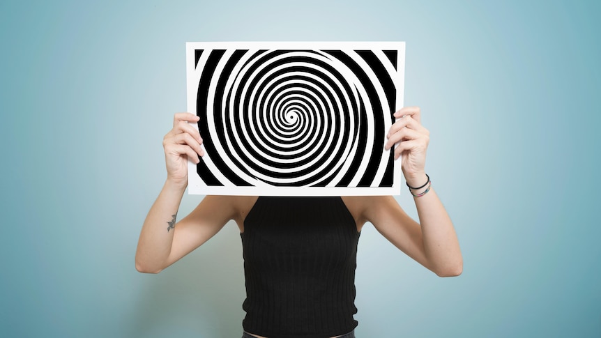 Person standing in front of a blue background, holding an artwork over their face of a black and white swirl 
