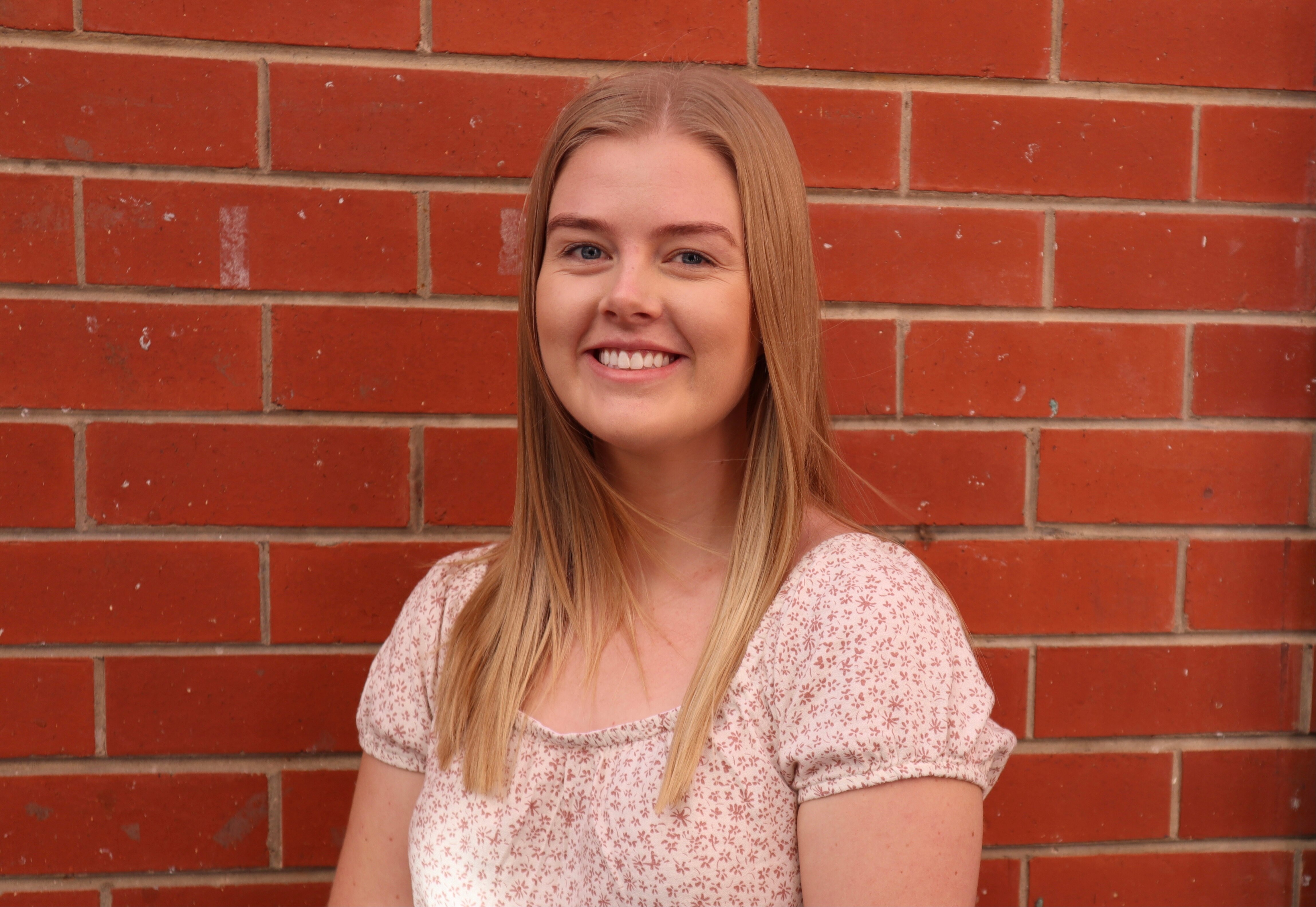 A young, blonde woman in a floral top in front of a red brick wall smiling