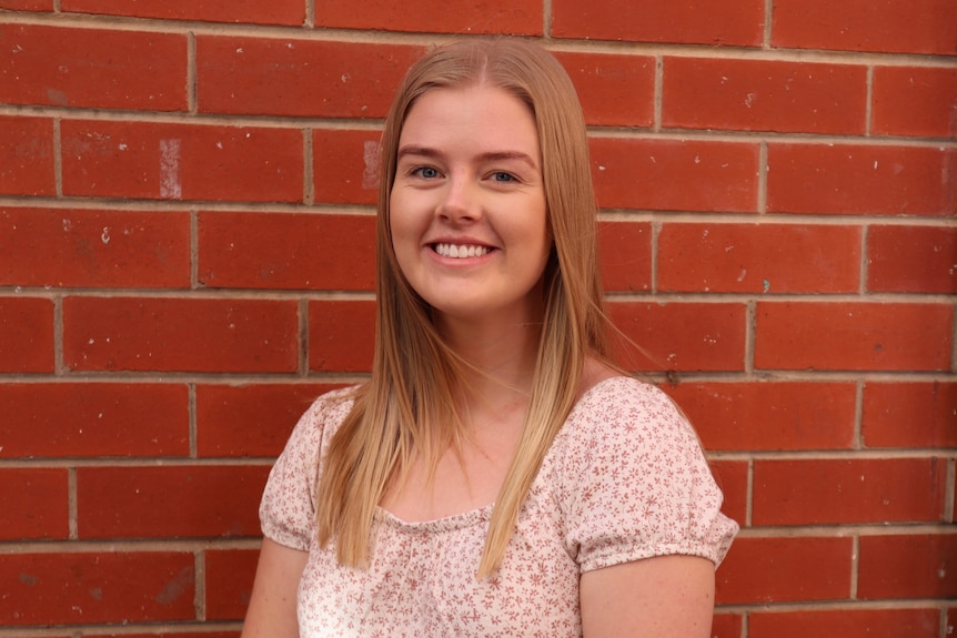 A young, blonde woman in a floral top in front of a red brick wall smiling