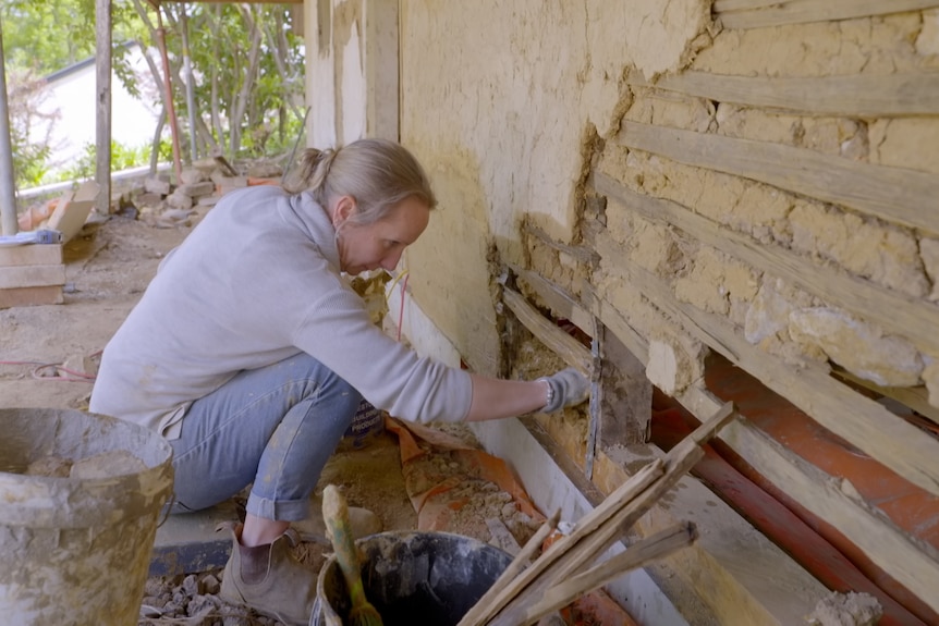 A woman sits at the front of an old mud cottage and rebuilds missing sections of it using her hands