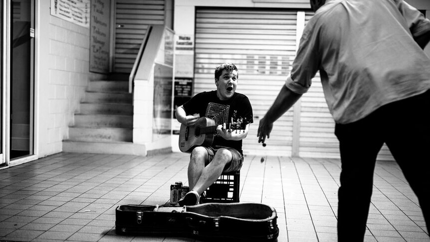 Young man with a guitar sitting on a milk crate busking in a mall