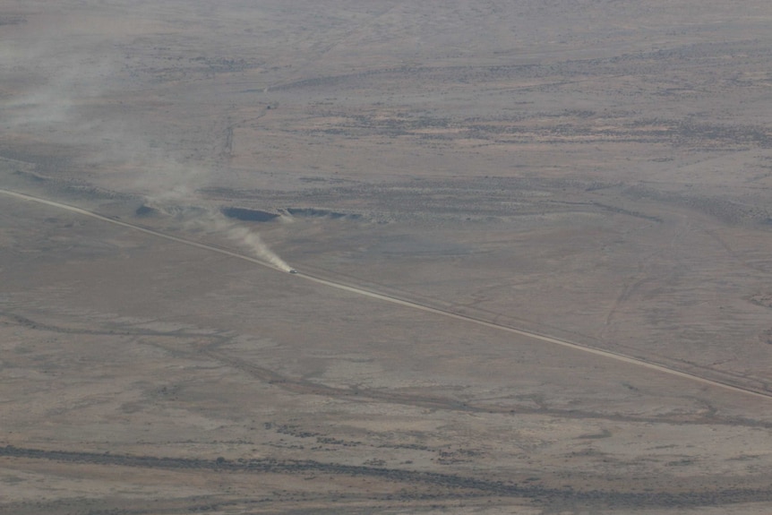 Wide aerial shot of a vehicle travelling along a dirt track through the desert.