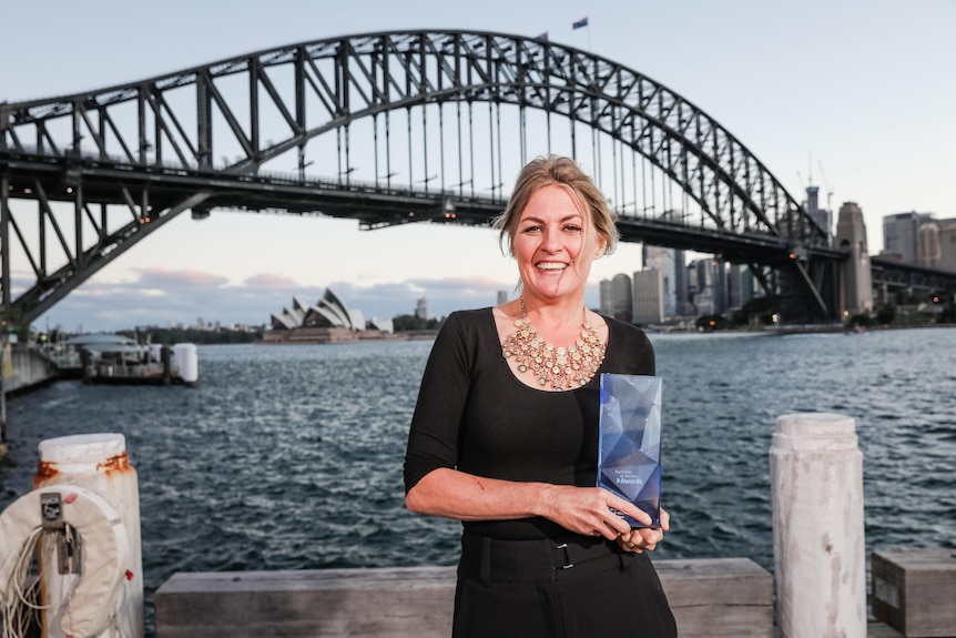 A woman with blond hair stands holding a trophy in front of the Sydney Harbour Bridge.