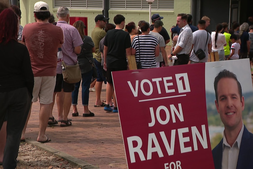 Casually dressed men and women queue behind a corflute for mayor candidate Jon Raven, says vote 1 beside photo.