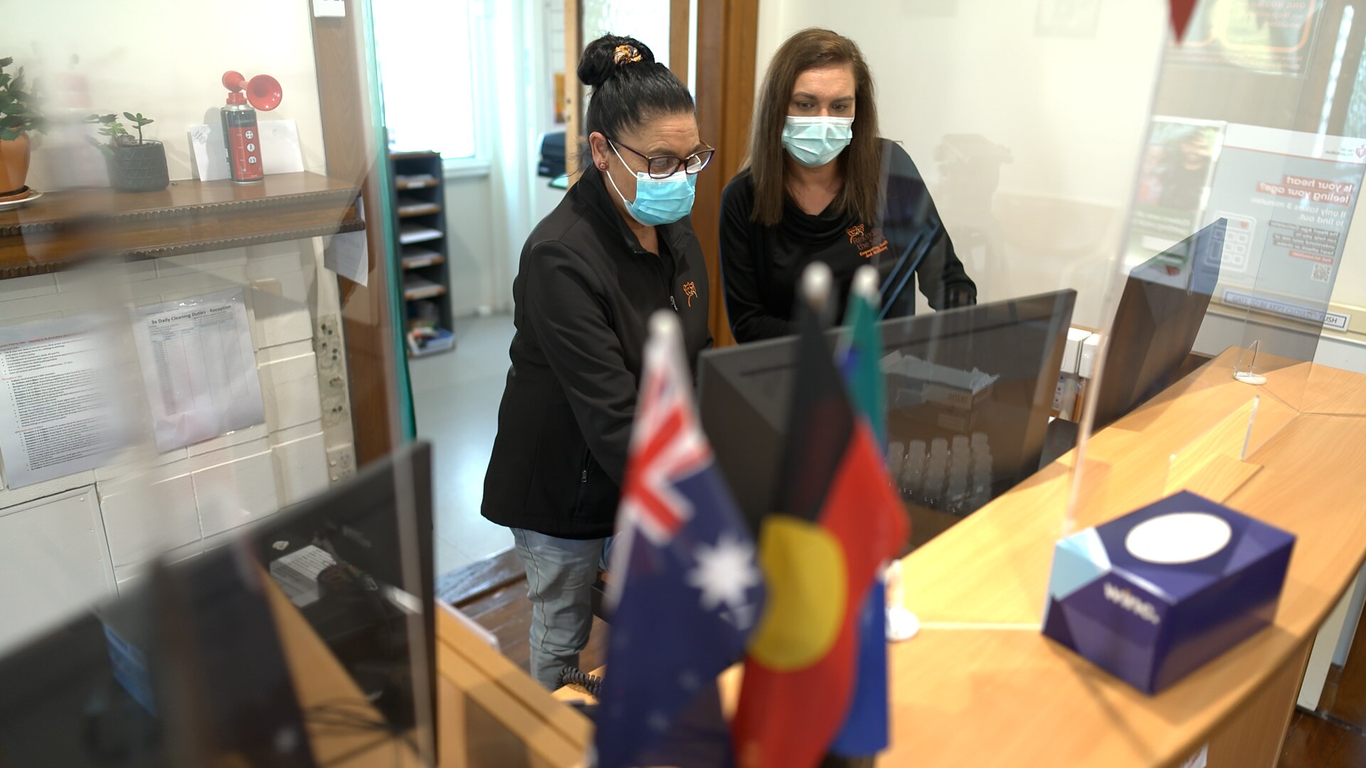 Two women, both wearing masks, at a reception desk with Australian and Aboriginal flags in the foreground.