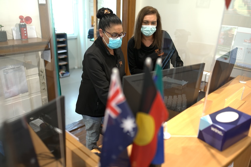 Two women, both wearing masks, at a reception desk with Australian and Aboriginal flags in the foreground.
